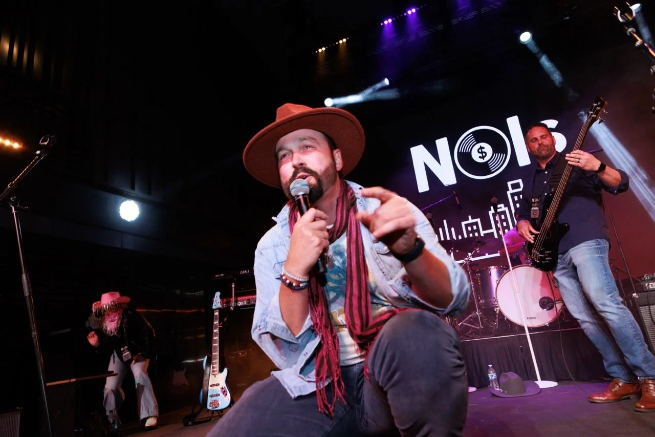 Performer with a microphone on stage, wearing a wide-brimmed hat, denim jacket, and scarf, with his hand gesturing toward the audience. In the background, another musician plays a bass guitar, and a woman with pink hair and a cowboy hat dances. The s
