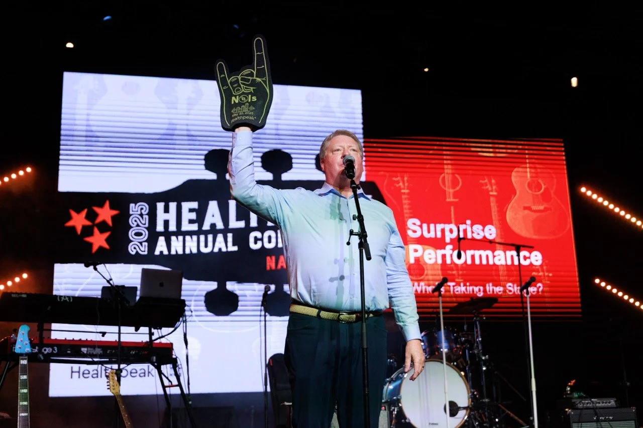 A man in a light blue shirt and dark pants is speaking into a microphone on stage during a conference. He is wearing a foam finger glove with the text 'NO!'. The background features a large screen displaying '2025 Healthy Annual Conference' and 'Surp