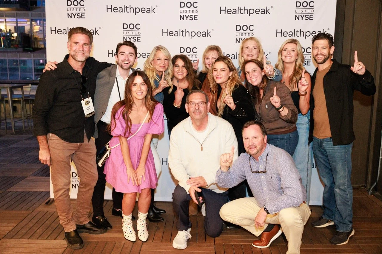 A group of 15 people posing for a photo at a Healthpeak event, standing in front of a backdrop with the company's name and NYSE listing. The group includes men and women, some making gestures with their hands, smiling and looking at the camera. It ap