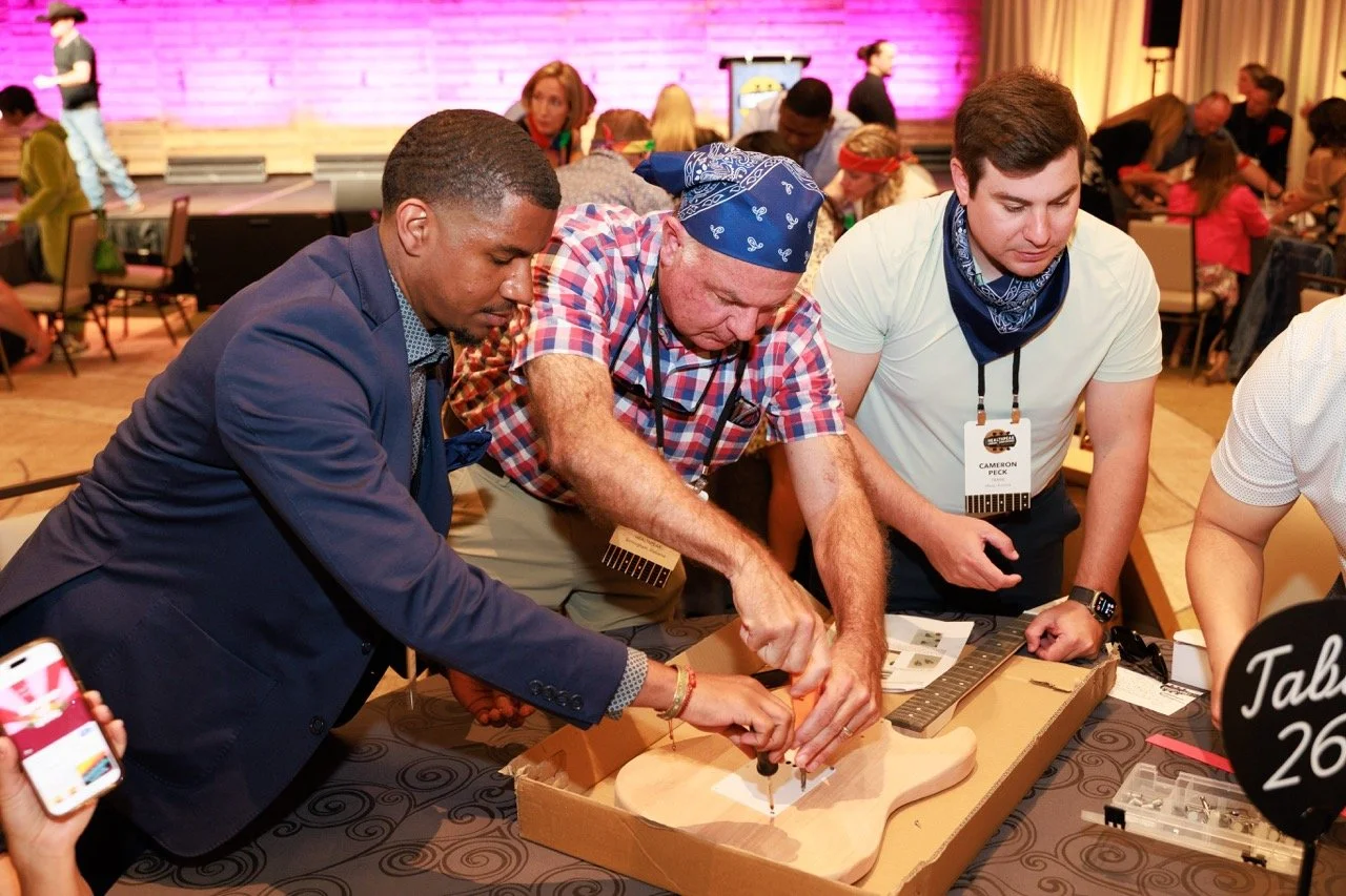 Three men are working on assembling a guitar at a table during an event, with a crowd of people in the background.