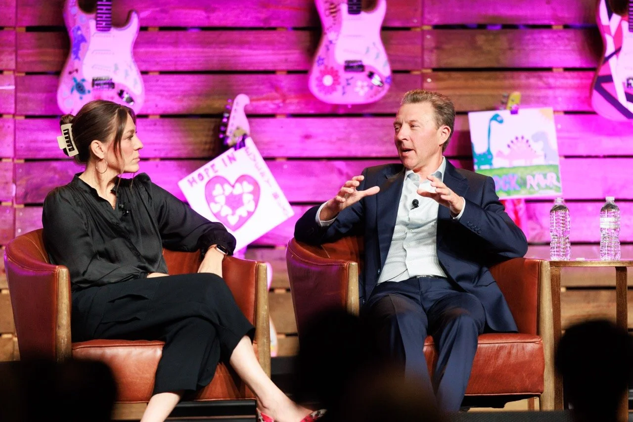 A woman and a man seated on stage, having a discussion. The woman has long hair in a bun with a large hair clip, wearing a black outfit. The man is gesturing with his hands, wearing a dark suit and white shirt. The stage background has pink guitars a