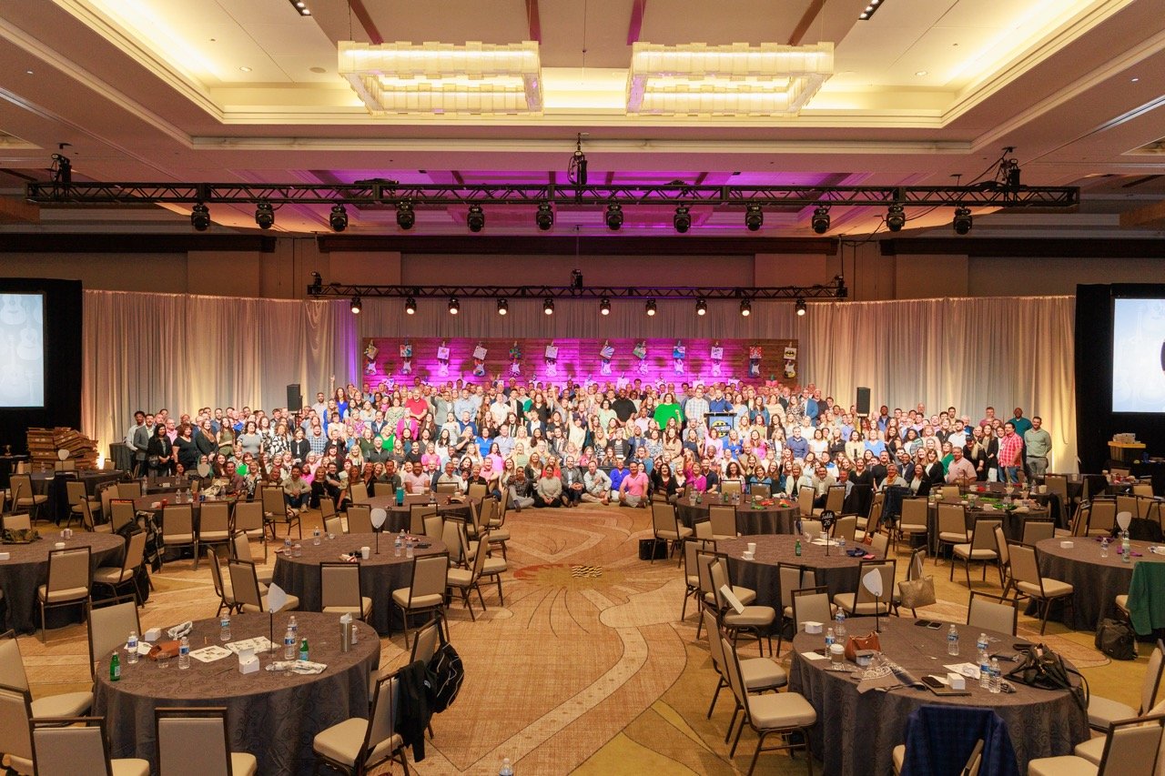Large banquet hall with many round tables and chairs, a stage filled with a large group of people, colorful lighting, and decorated backdrop for an event or celebration.