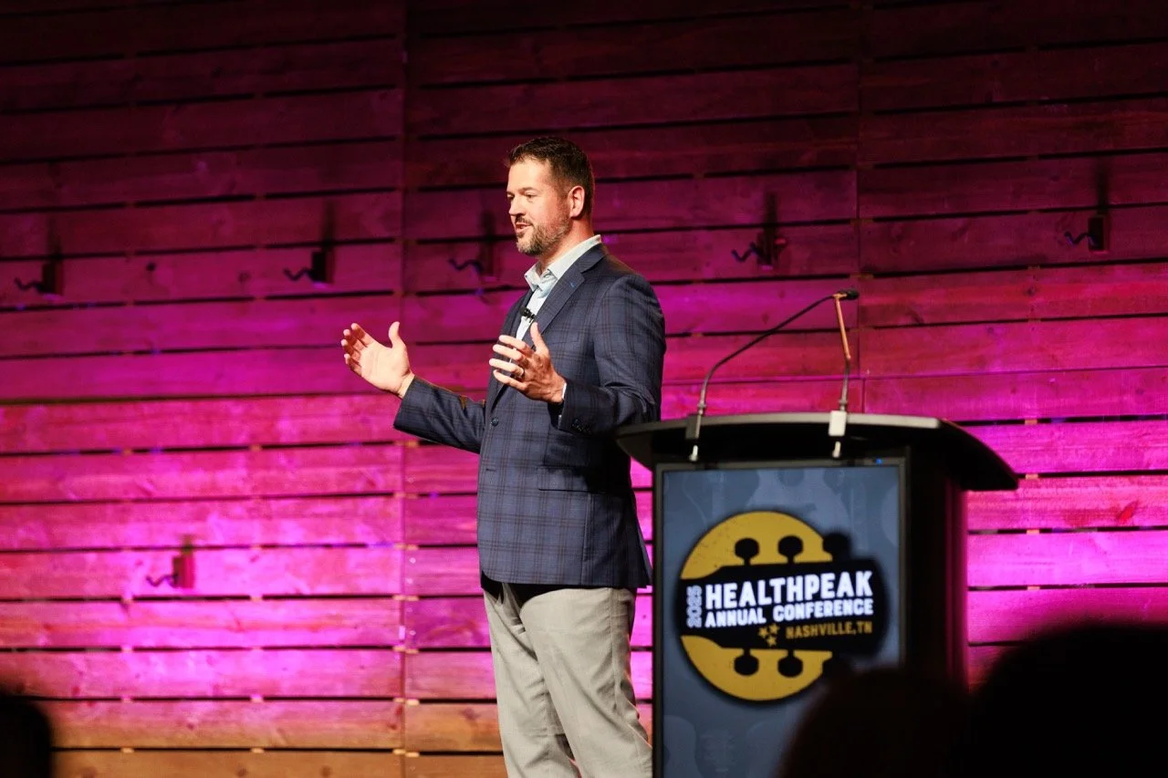A man in a plaid blazer and beige pants giving a presentation at the 2023 Healthpeak Annual Conference in Nashville, Tennessee. He stands on a stage with a pink-lit wooden backdrop.