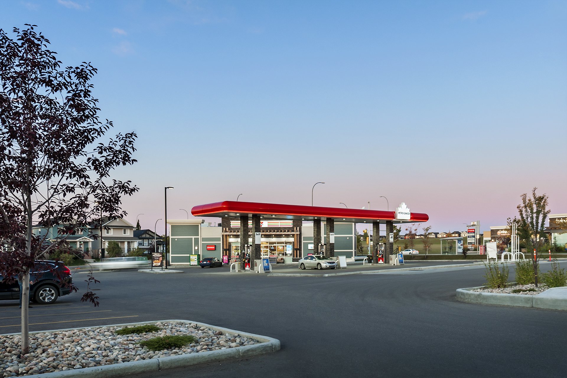A Shell gas station at dusk with a parking lot in the foreground, trees, residential homes, and a clear sky in the background.