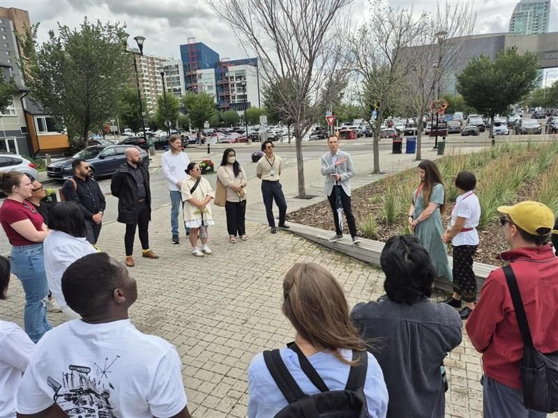 Group of people gathered outdoors on a paved area near trees, listening to a woman in traditional dress, with city buildings and parked cars in the background.