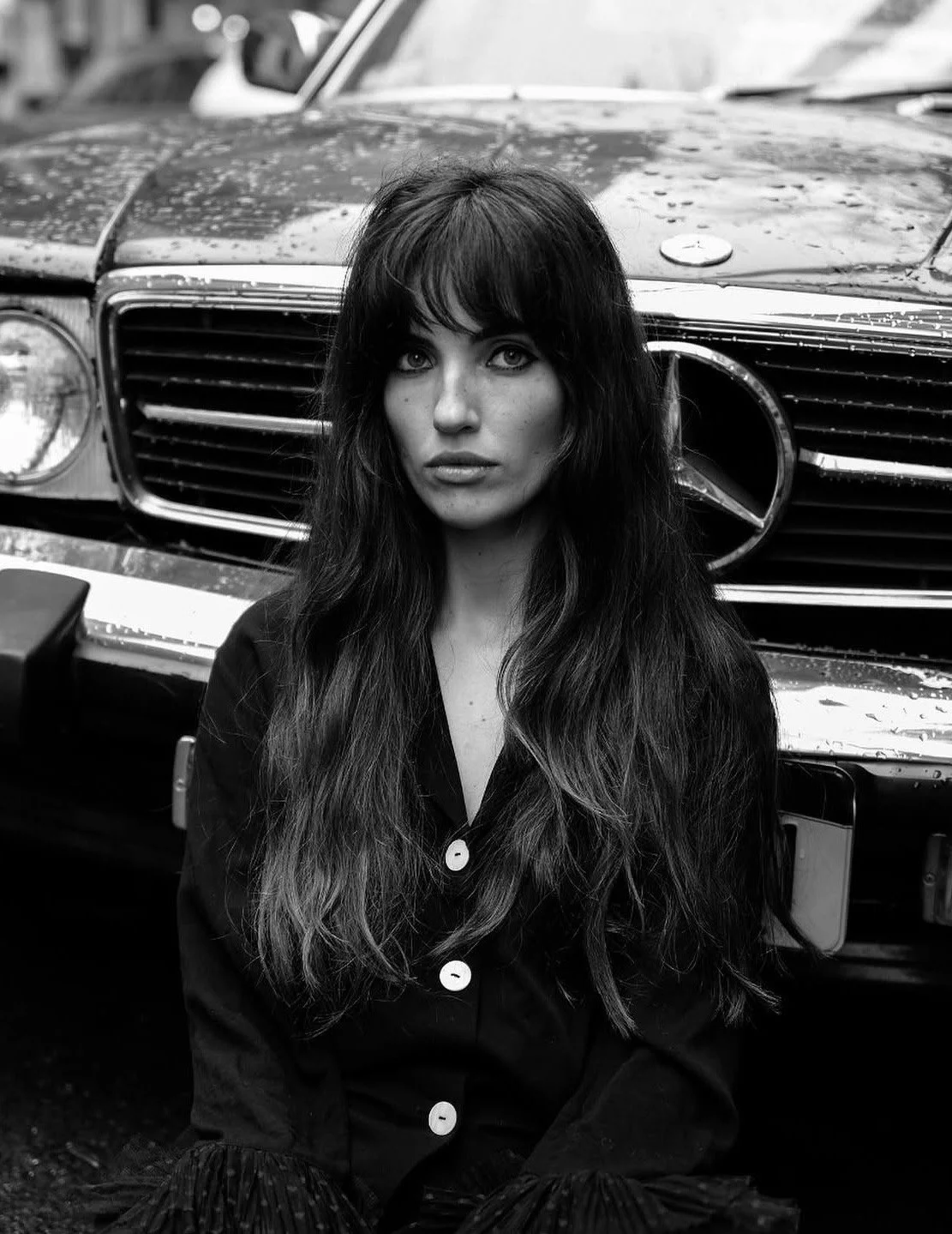 A woman with long dark hair, wearing a button-up shirt, sitting in front of a wet vintage Mercedes-Benz car, in black and white.
