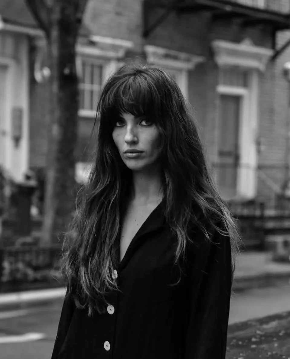 Black and white photo of a woman with long, wavy hair and bangs, wearing a dark button-up shirt, standing outdoors in an urban setting with buildings and a car in the background.