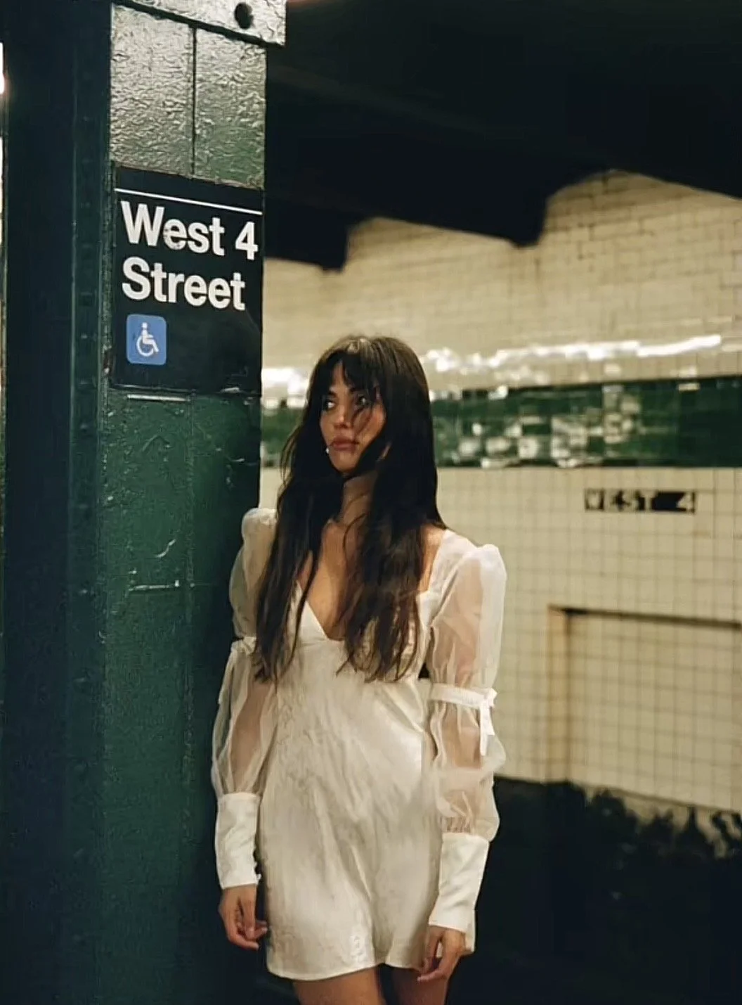 A woman with long dark hair wearing a white dress with sheer puffed sleeves, standing against a green wall with a sign that reads "West 4 Street" and a wheelchair accessibility symbol, in an underground subway station.