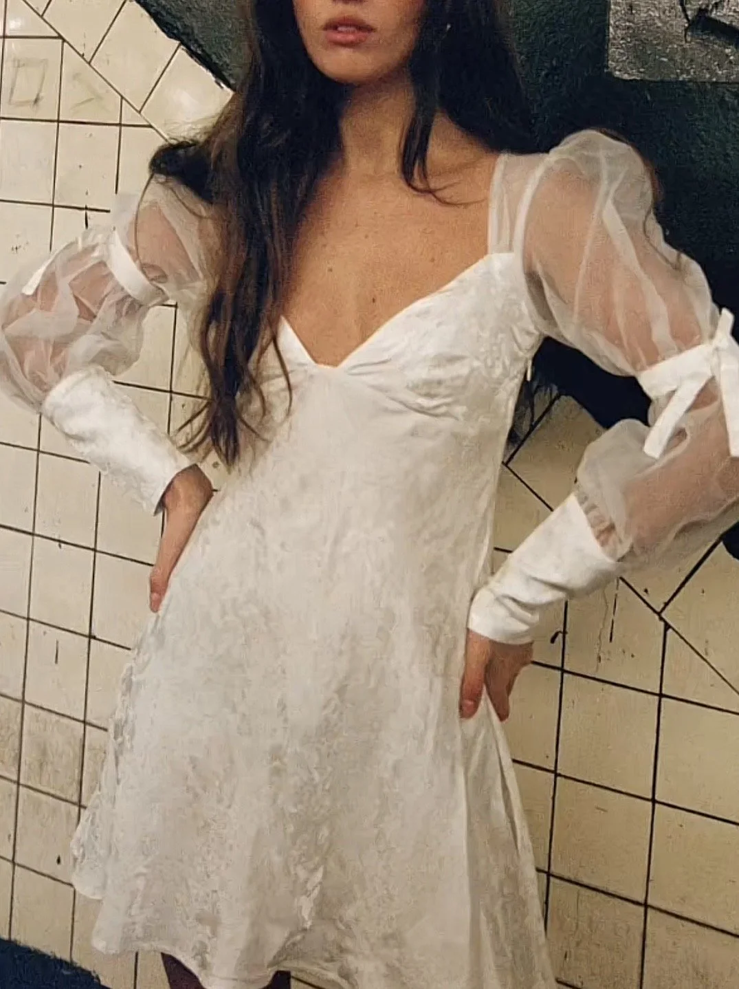A woman wearing a white dress with sheer puffed sleeves, standing against a tiled wall.