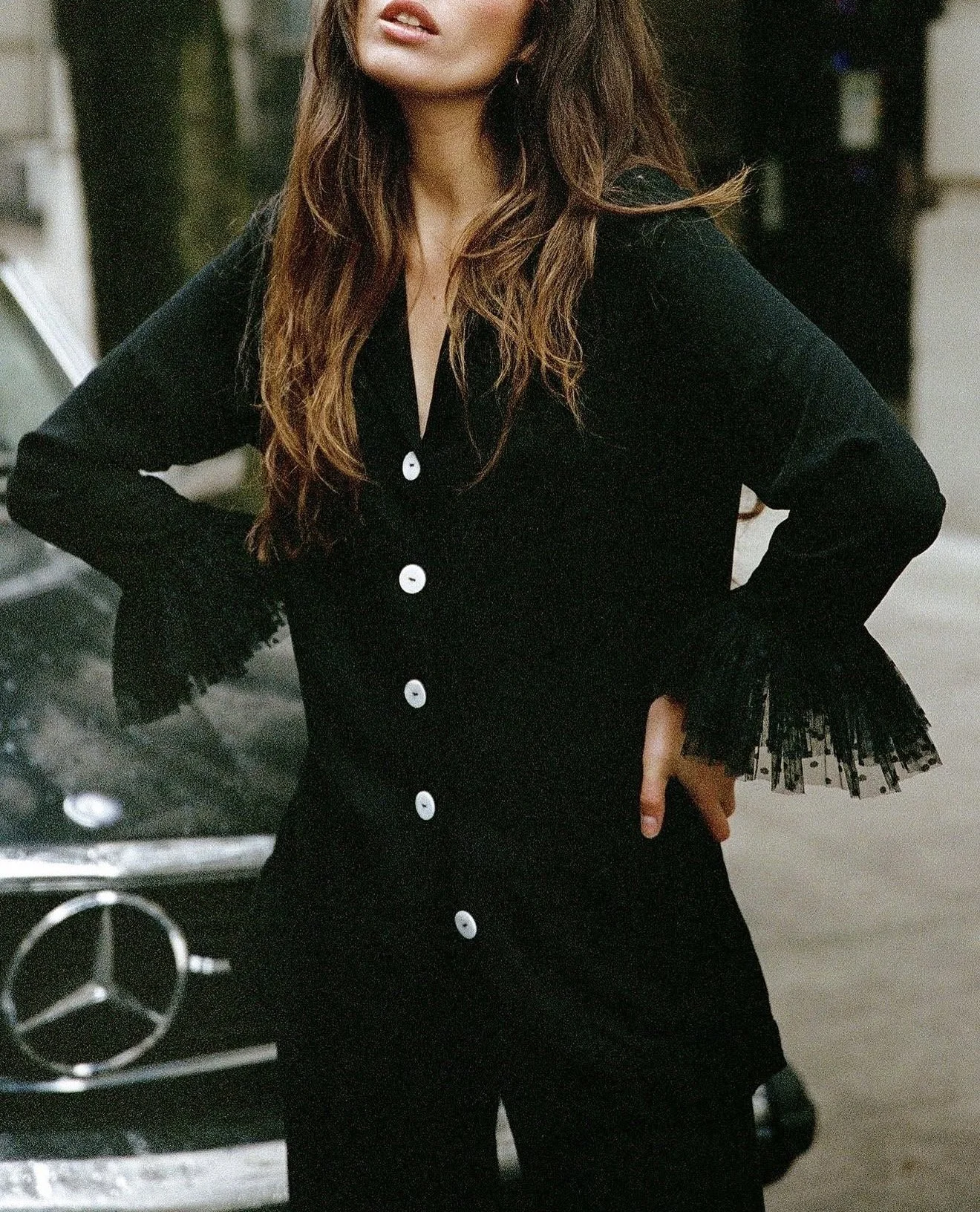 A woman with long wavy brown hair wearing a black dress with white buttons and lace cuff details, standing in front of a car with a Mercedes-Benz logo, in an indoor parking garage.
