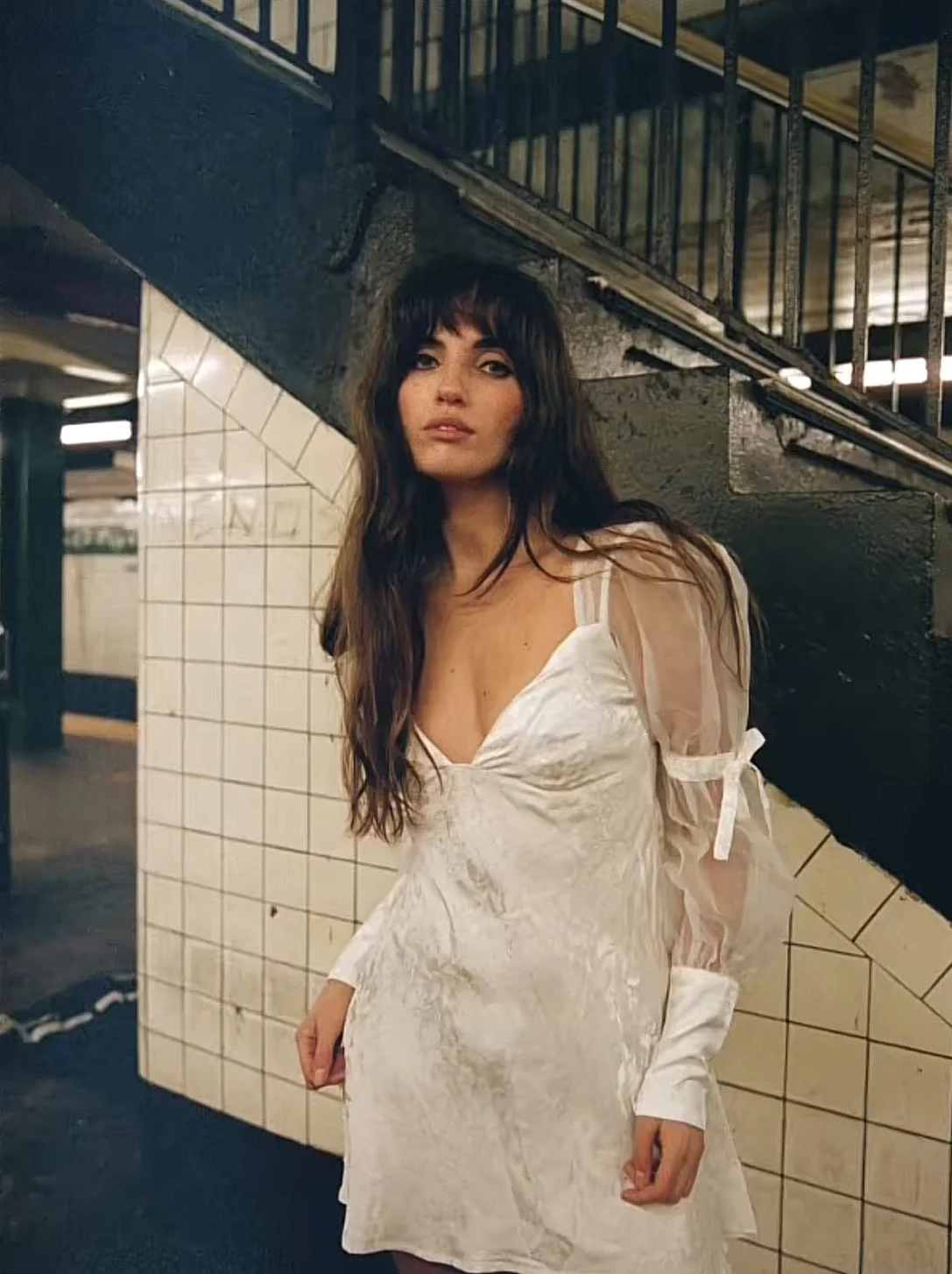 A woman in a white dress with lace and sheer sleeves stands under a staircase in a subway station with tiled walls.
