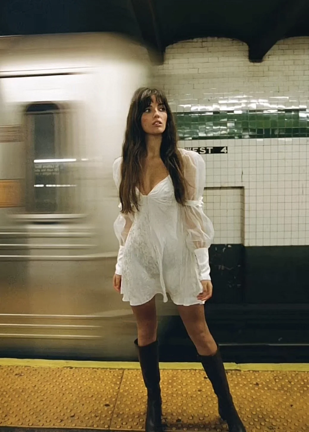 A woman in a white dress with long sleeves and black knee-high boots stands on a subway platform as a train passes by behind her.