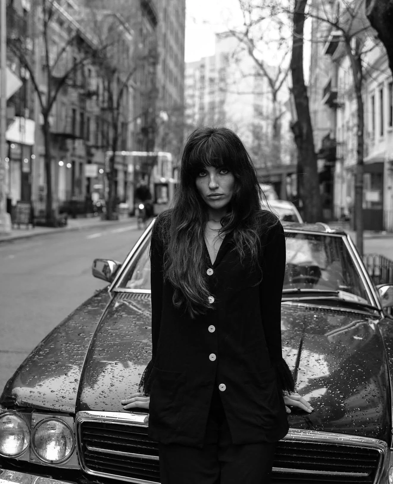 A woman with long dark hair and bangs standing in front of a vintage car, on a city street with buildings and leafless trees in the background, black and white photo.