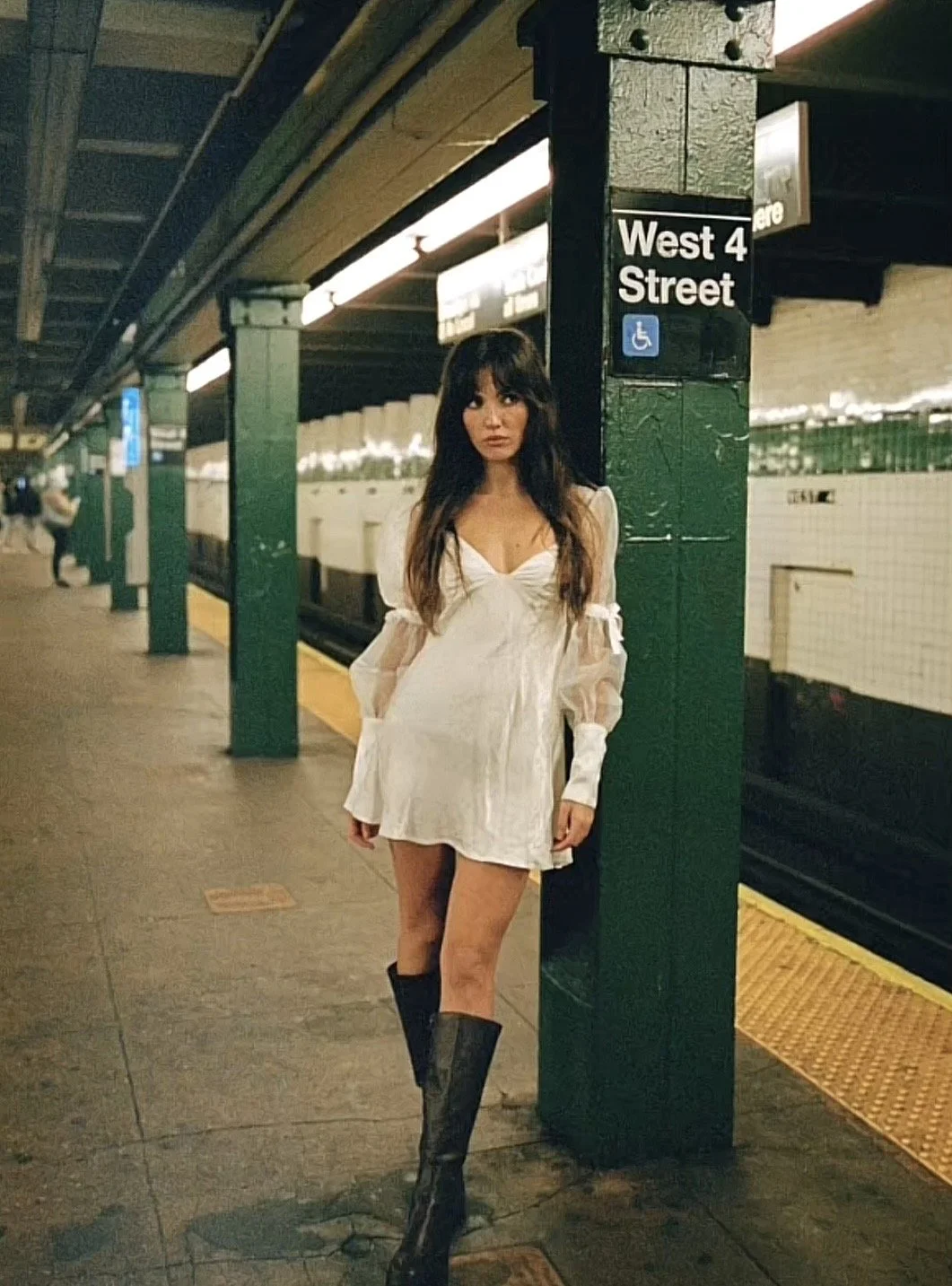 A woman in a white dress and tall black boots standing on a subway platform at West 4th Street station. The platform has green columns and a yellow safety strip along the edge.