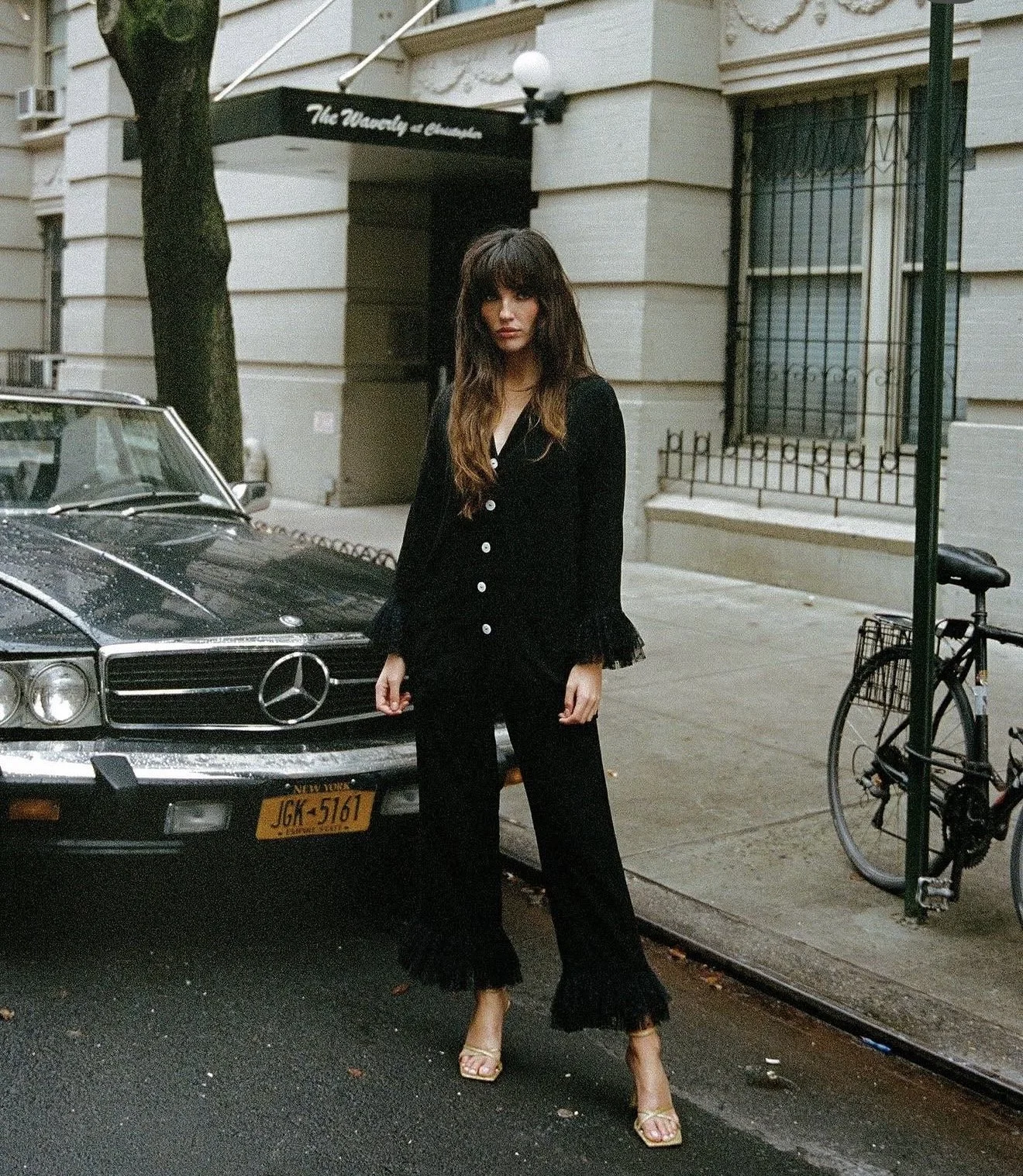 A woman in a black outfit with lace details stands on a city street next to a black vintage Mercedes-Benz car with New York license plates. There is a bicycle locked to a nearby post, and a building with barred windows in the background.