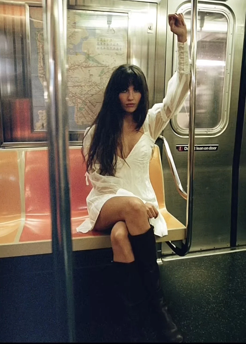 A woman sitting on a subway seat with her left arm raised holding onto a pole, wearing a white dress and black boots, with long dark hair and bangs.