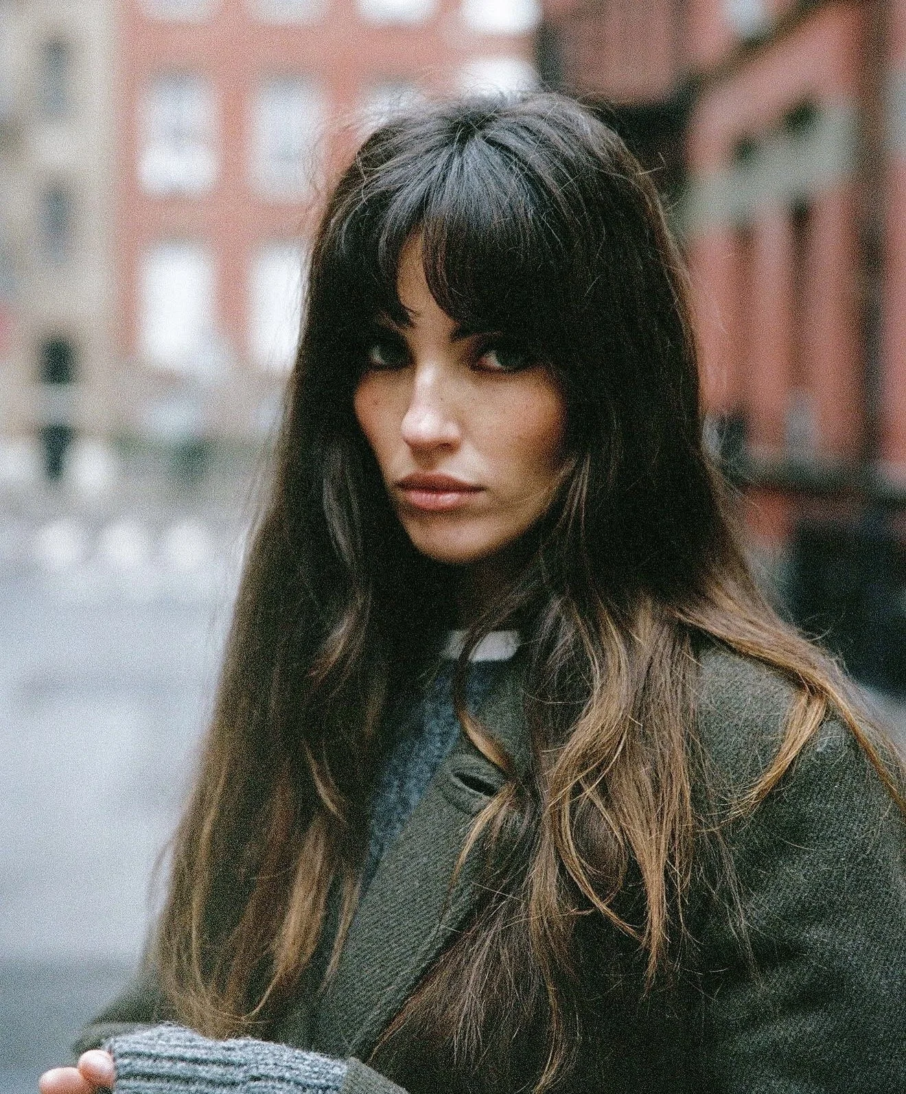 A woman with long wavy hair and dark hair, wearing a dark jacket, standing outdoors in an urban area with blurred buildings in the background.