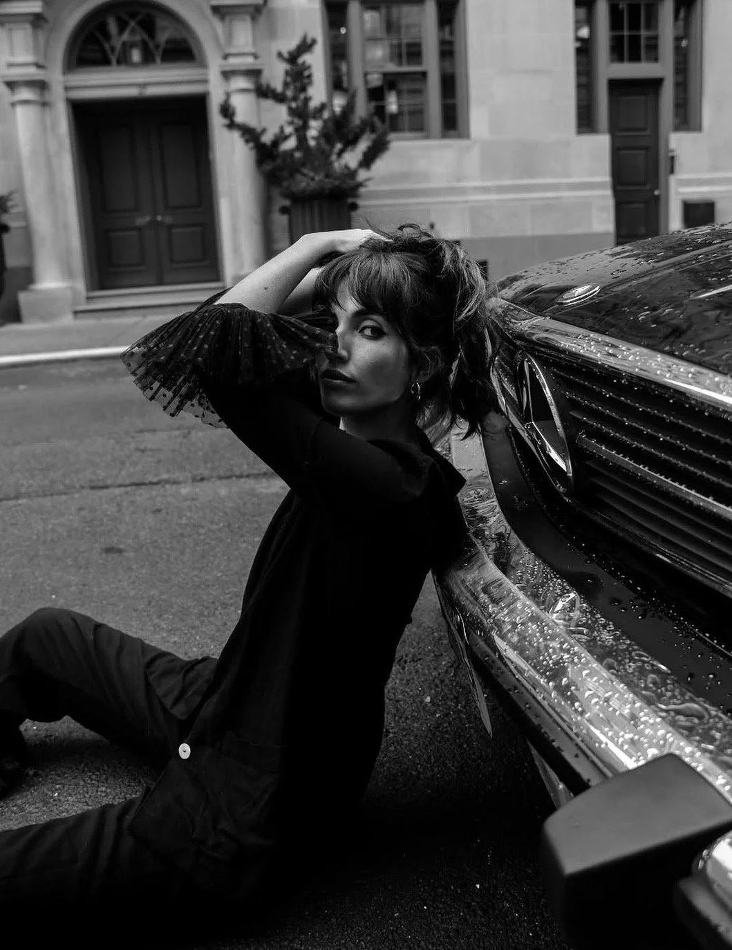 A woman with dark hair and a hand on her head leans against a wet luxury car on a city street in black and white.