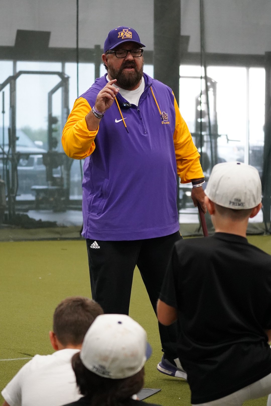 A man wearing glasses, a purple and yellow sports jacket, and a purple baseball cap with ESPN logos is speaking to a group of children indoors. The children are sitting on the floor, some wearing hats, listening attentively.