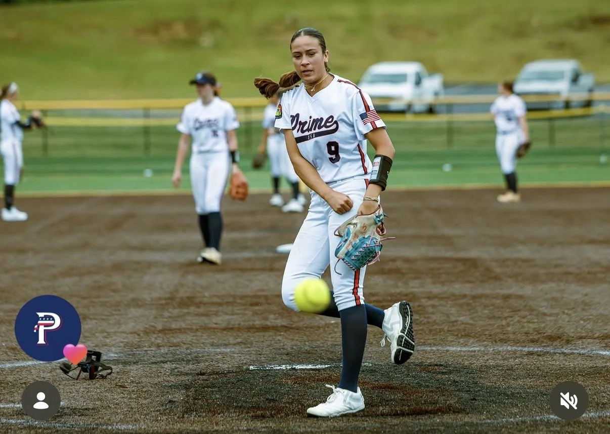 A female softball player wearing a white uniform with red and navy accents, practicing pitching on a softball field. She is in a follow-through pose after throwing a yellow softball, wearing a baseball glove, and additional players are in the background on the field.