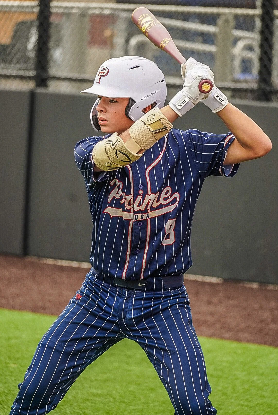 A young male baseball player in a navy blue pinstriped uniform with red and white accents, holding a bat ready to swing, standing on a baseball field with a batting helmet and gloves.