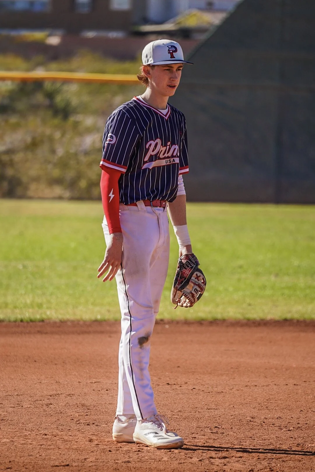 A young male baseball player standing on the field, wearing a navy pinstripe jersey that says 'Prime USA,' white pants with a dirt stain, a cap with a 'P' logo, red and white sleeves, and holding a glove in his left hand.