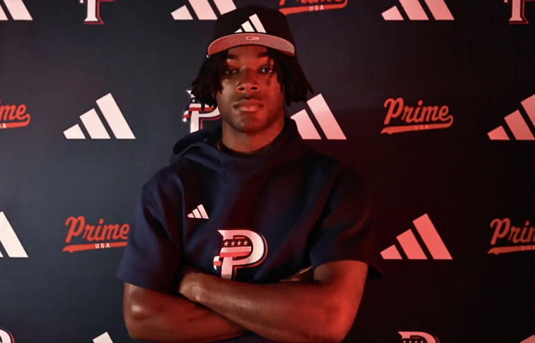 Young man with dreadlocks wearing a black hoodie, baseball cap, and posing with arms crossed in front of a backdrop with Adidas and Prime logos.