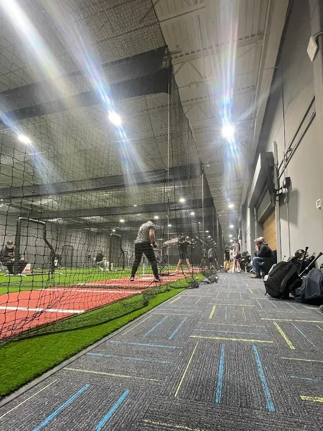 An indoor golf practice area with a netted hitting zone, artificial turf, and multiple people practicing or sitting along the side.