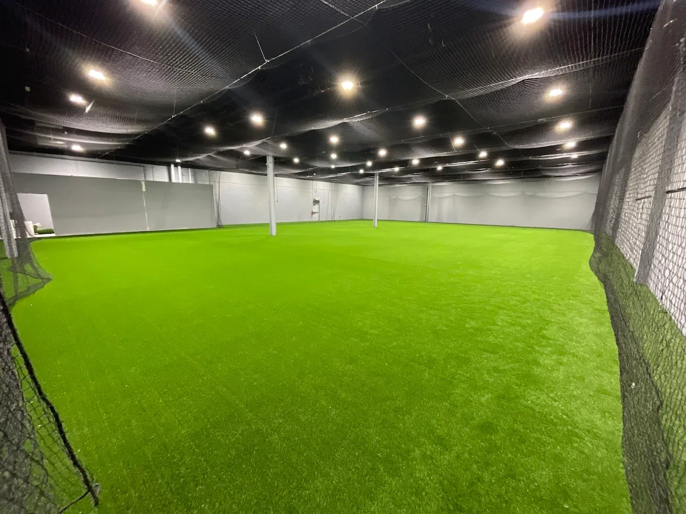 Indoor sports facility with bright green artificial turf, surrounded by black netting, and illuminated by ceiling lights.
