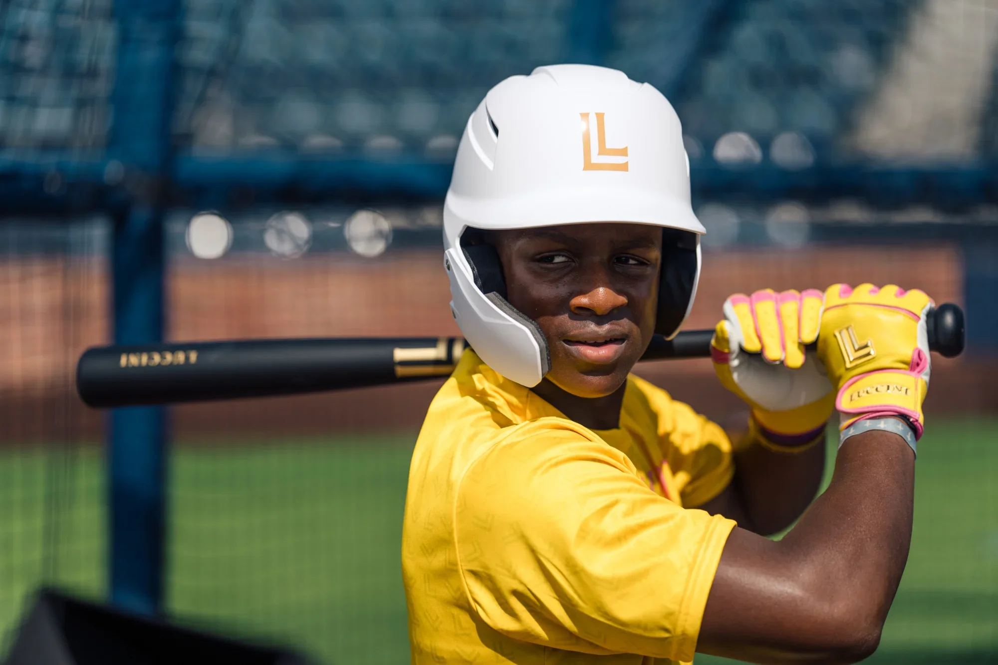 A young baseball player in a yellow jersey and matching gloves holds a bat, wearing a white helmet with a gold logo, at a baseball field.