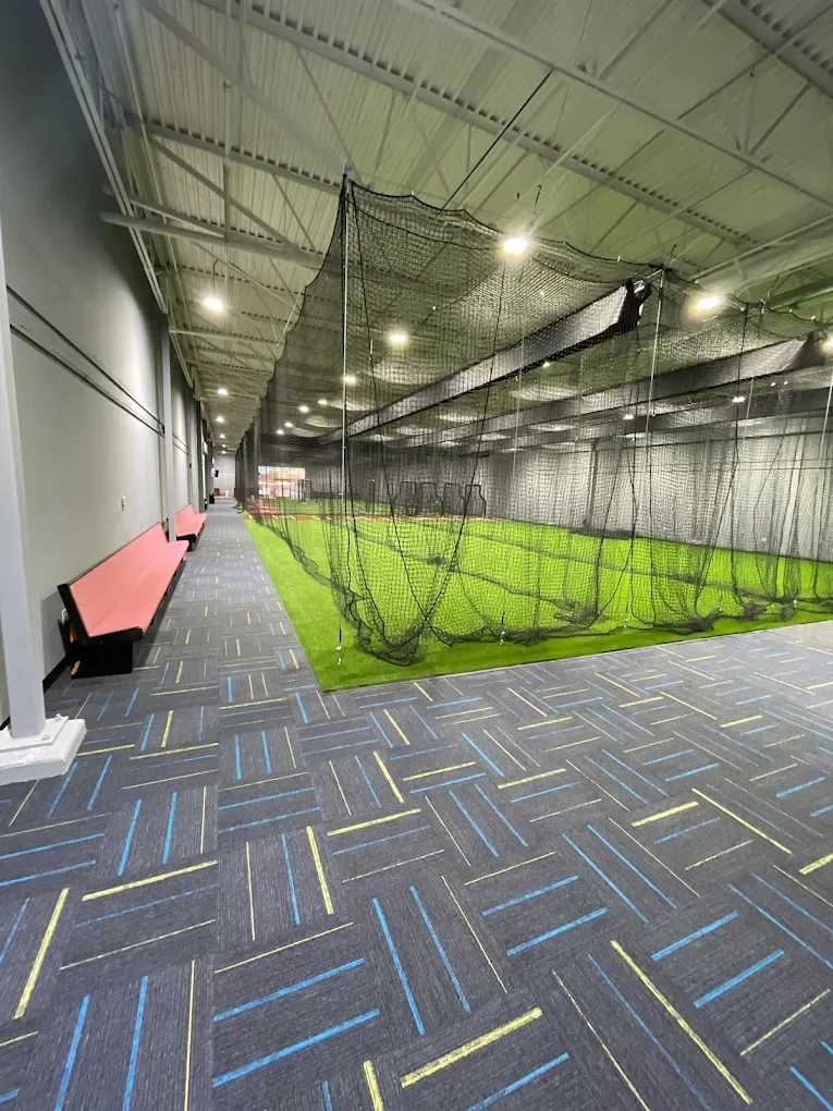 Indoor golf practice area with artificial turf and a black net for practicing swing shots, located inside a building with colorful striped carpet and pink benches along the wall.