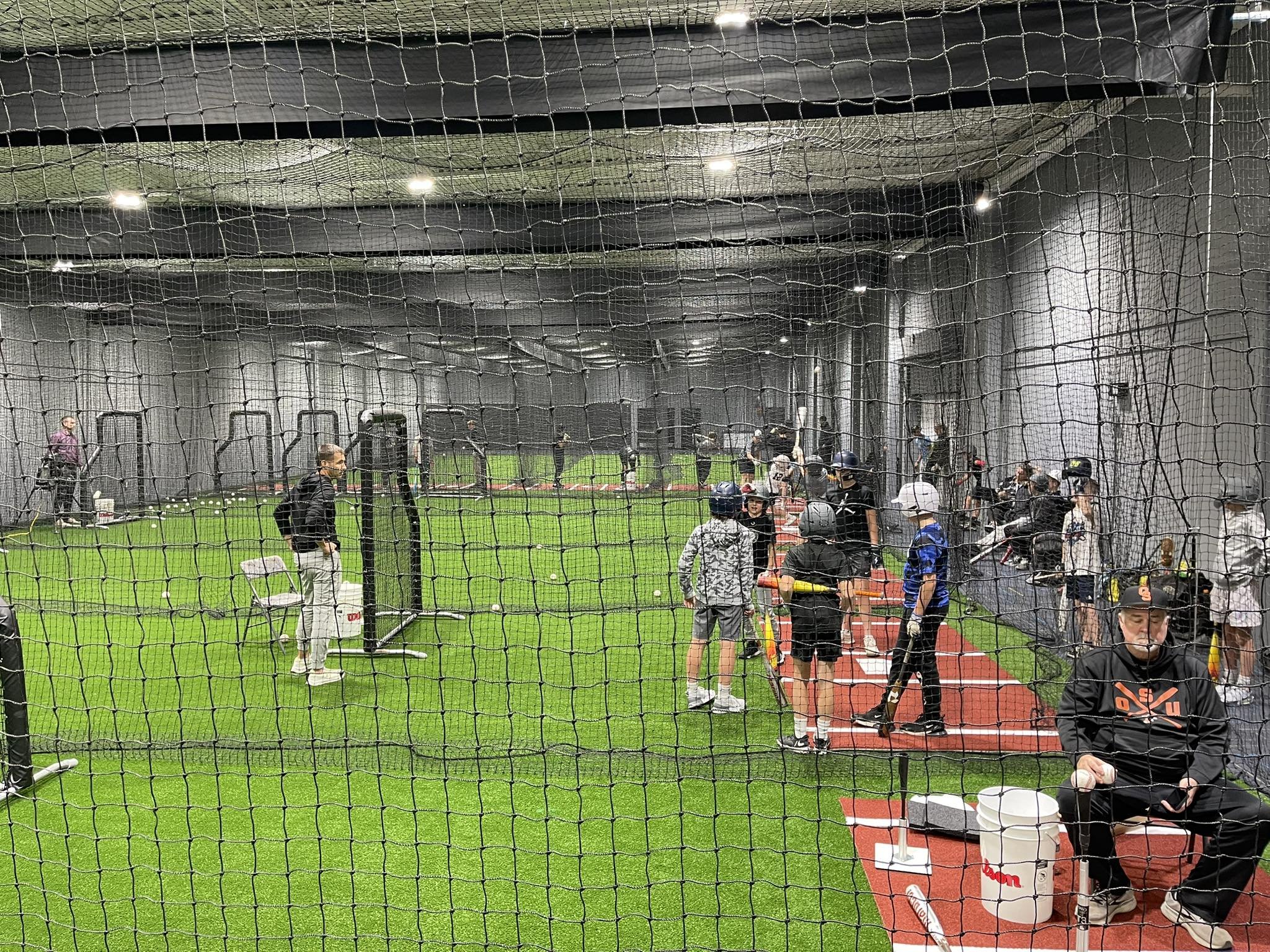 Indoor baseball or softball training facility with players and coaches, view through protective netting.