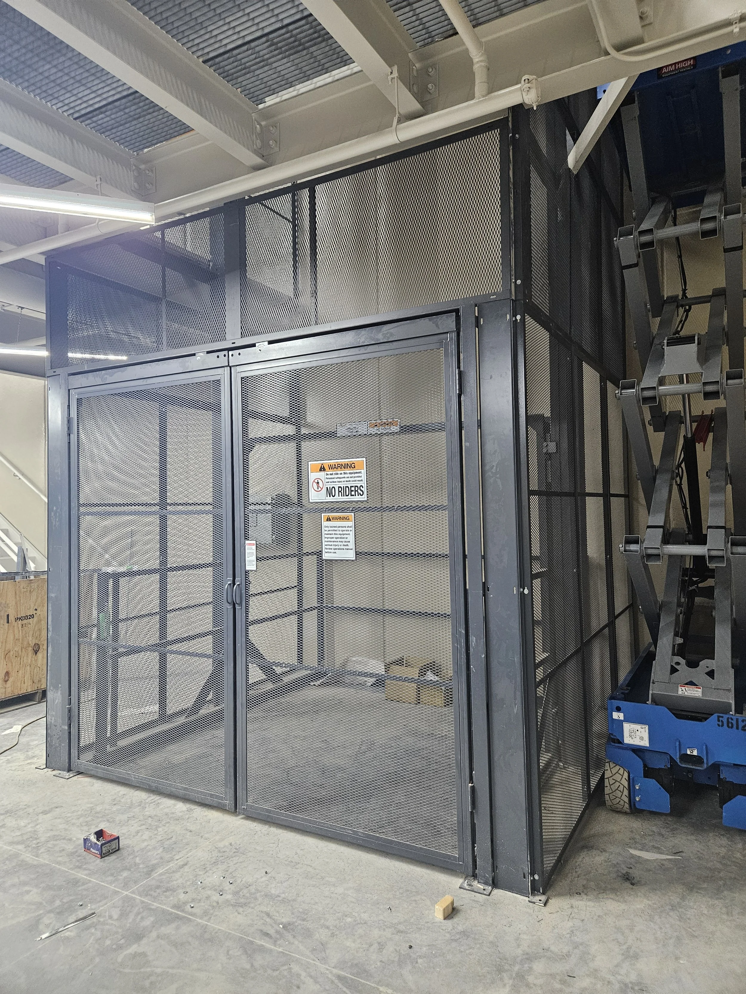 Construction site with a metal cage for an elevator, safety signs on the door, and construction tools and objects on the floor.