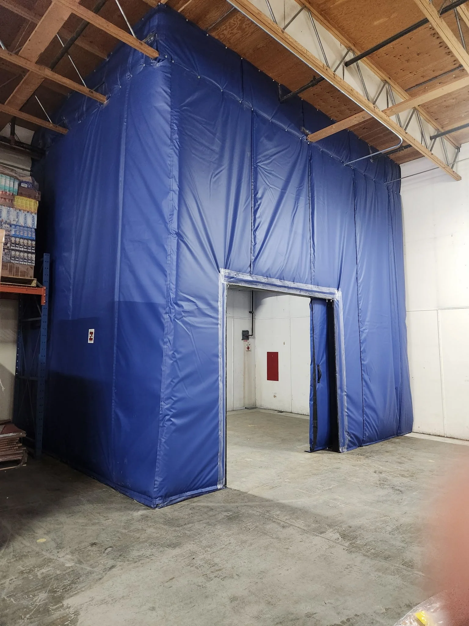 A blue fabric temporary structure or curtain inside a warehouse with wooden ceiling and concrete floor.
