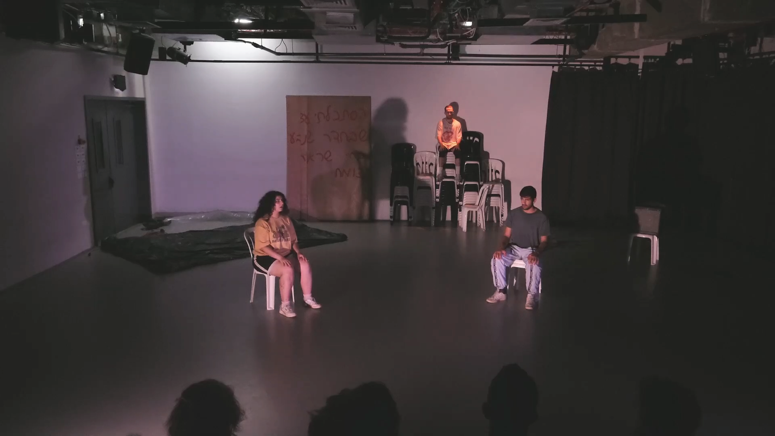 Three people are in a room with minimal props. One person sits on a white chair to the left, another sits on a white chair to the right, and the third stands on a stack of chairs at the center back. There is a brown board with writing on it and a black cloth on the floor in the background, with stage equipment and black curtains on the right side.