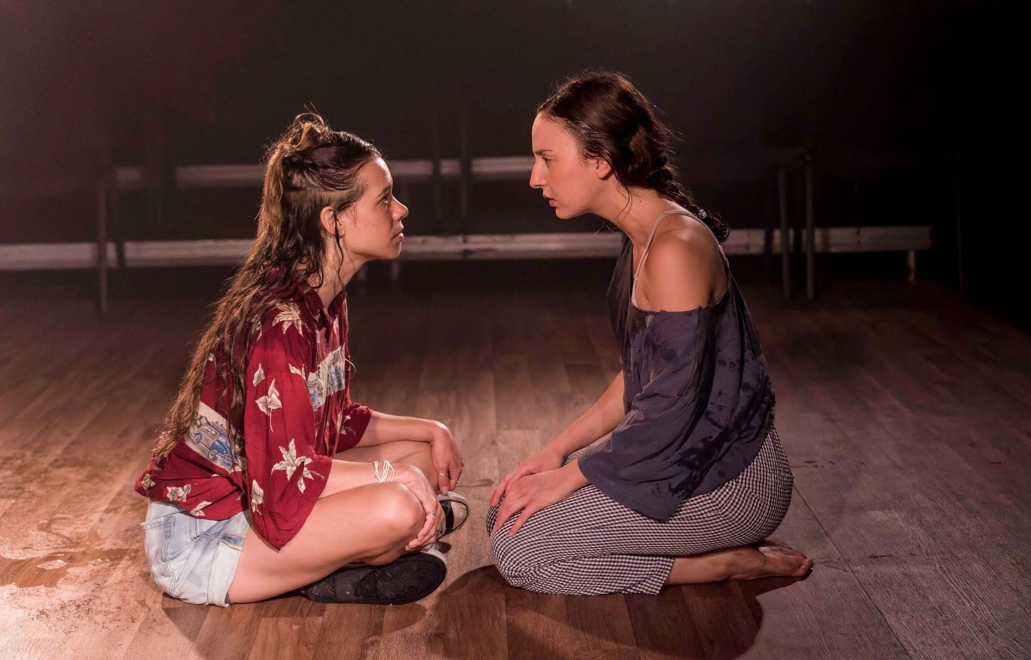 Two women with dark, curly hair and light skin are kneeling and holding hands in front of a dark background. They are facing each other with serious expressions, and are positioned close together, touching knees. The woman on the left is wearing a red patterned shirt and white shorts, and the woman on the right is dressed in a black off-shoulder top and checkered pants.