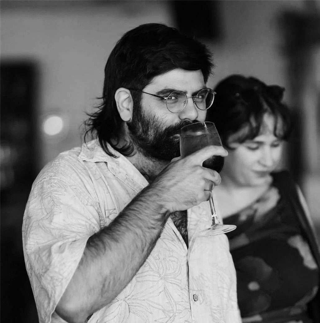 A man with dark hair, glasses, and a beard holding a glass of aperol spritz, with a woman in the background in an indoor setting.