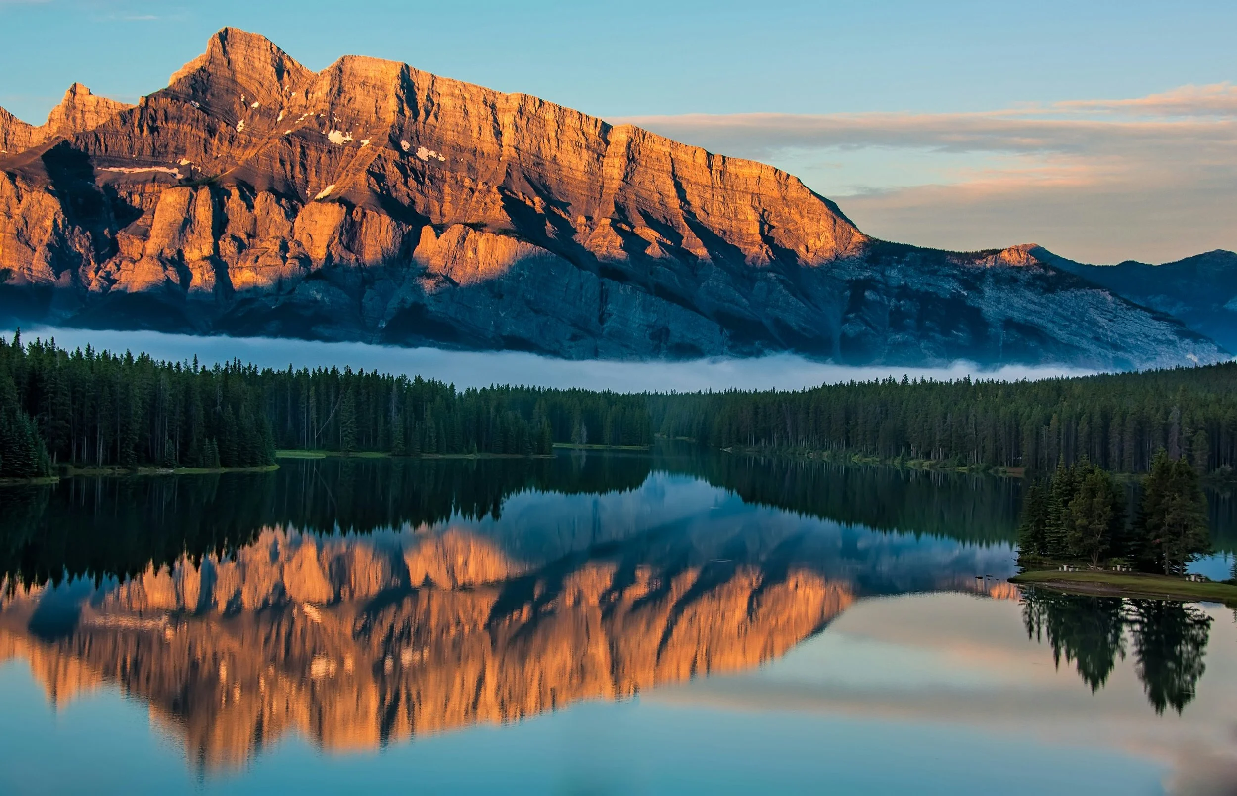 Sunset illuminating a rugged mountain range reflected in a calm lake surrounded by a dense forest.