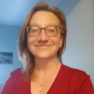 A woman with glasses and shoulder-length hair wearing a red top, smiling indoors.