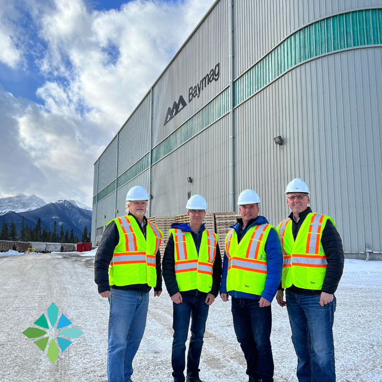 Four men in hard hats and high-visibility vests standing in front of a large industrial building labeled 'BayMag,' with snowy ground and mountains in the background.