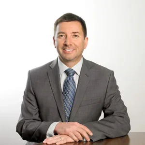 A man in a gray suit with a blue tie, smiling and sitting with his hands on a white surface, against a plain white background.