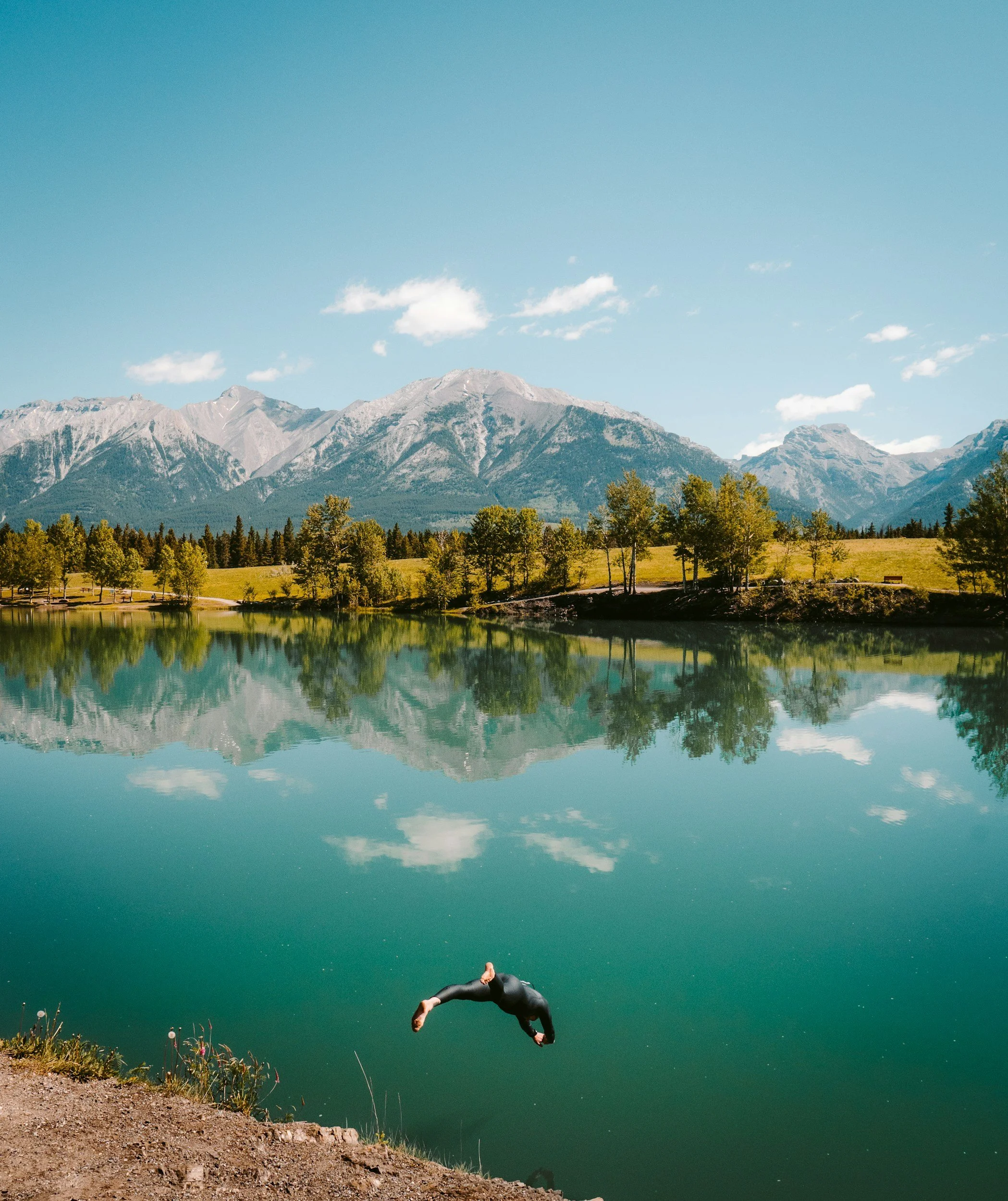 Person diving into calm green lake from rocky shoreline on sunny day with mountains and blue sky in background.