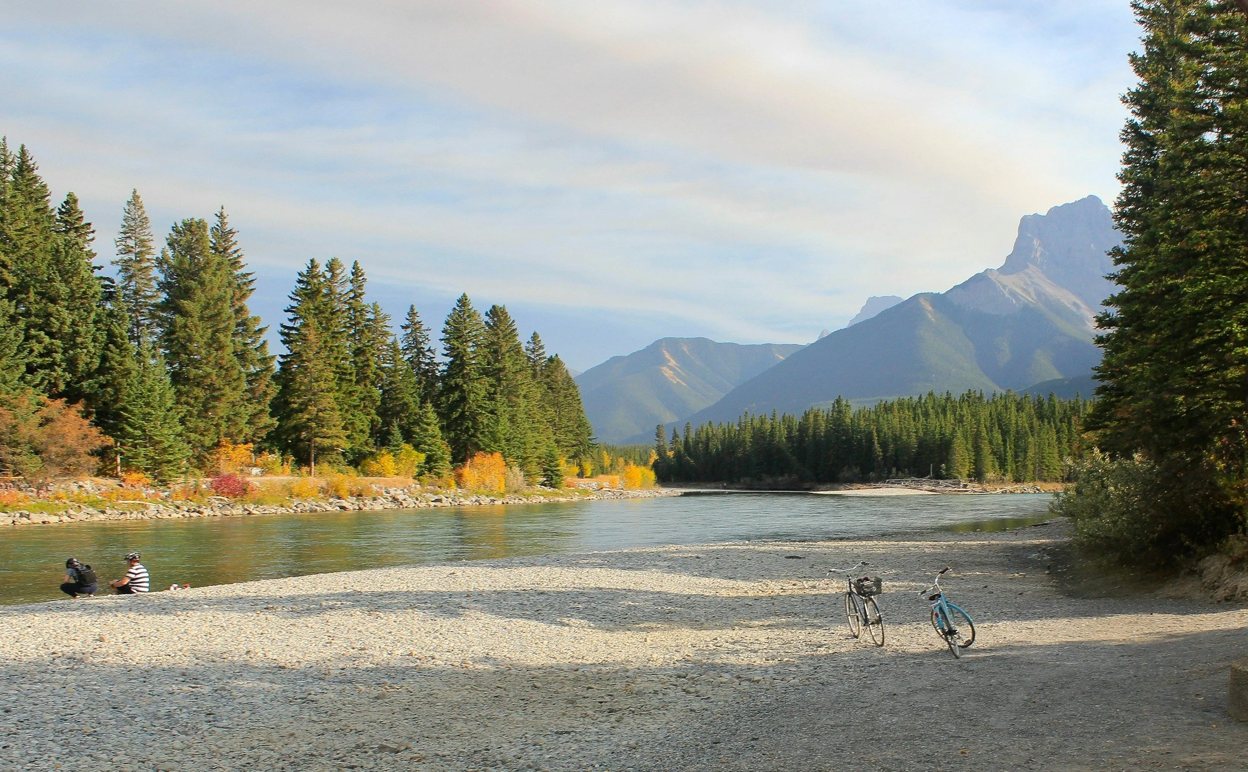 A river running through a forested area with mountains in the background, and two people sitting on the rocky shoreline near two bicycles.