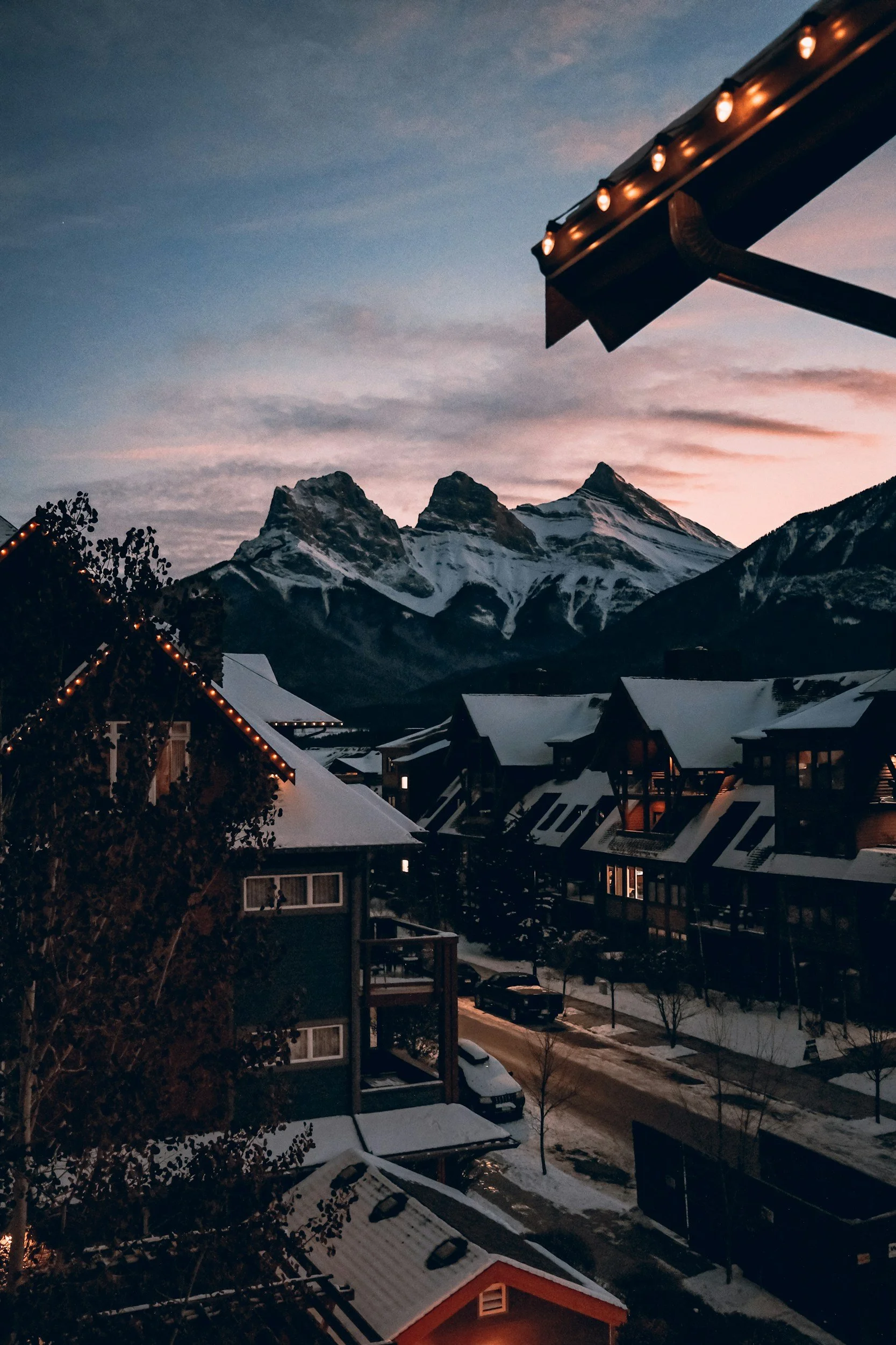 Snow-covered rooftops of houses in a mountain town, with tall snow-capped mountains in the background during dusk or dawn, and a partly cloudy sky with shades of pink and blue.