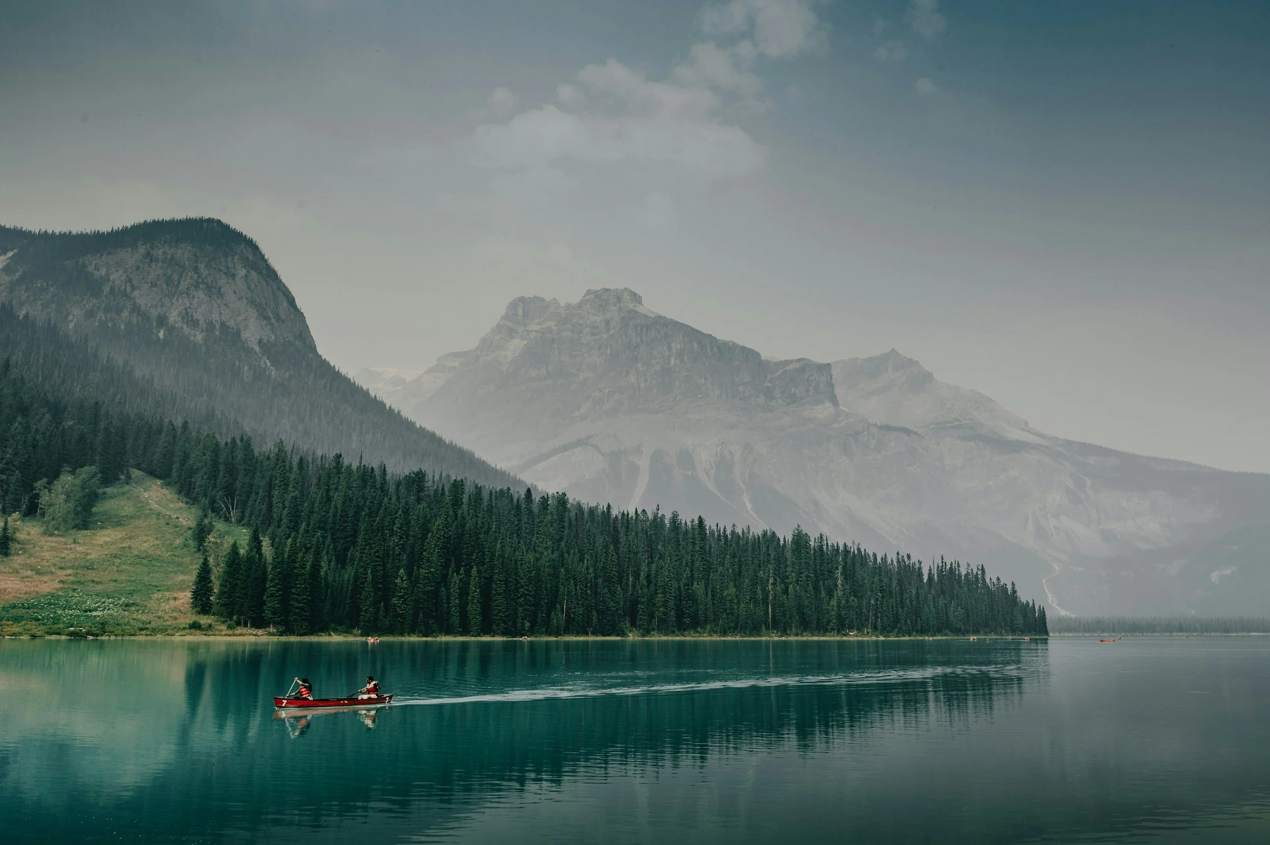 A lake surrounded by tall evergreen trees and towering mountains in the background, with two people in a red canoe paddling on the water.