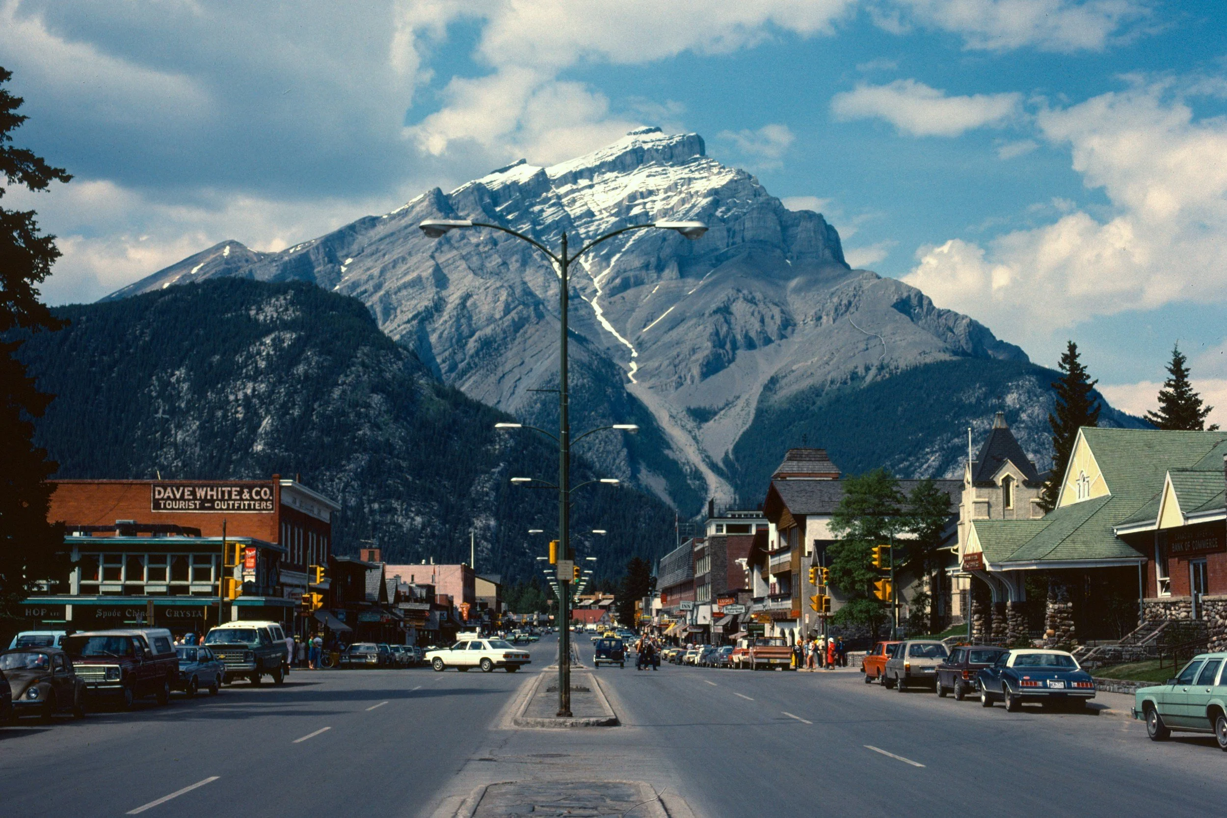 A small town street with parked cars, storefronts, and a crosswalk. In the background, there is a mountain with snow on its peak and a partly cloudy sky.