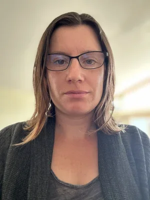 A woman with shoulder-length brown hair wearing glasses and a dark top, looking into the camera indoors.