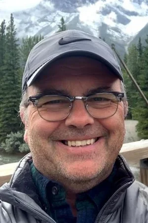 Smiling man wearing glasses and a cap outdoors with mountains and trees in the background.