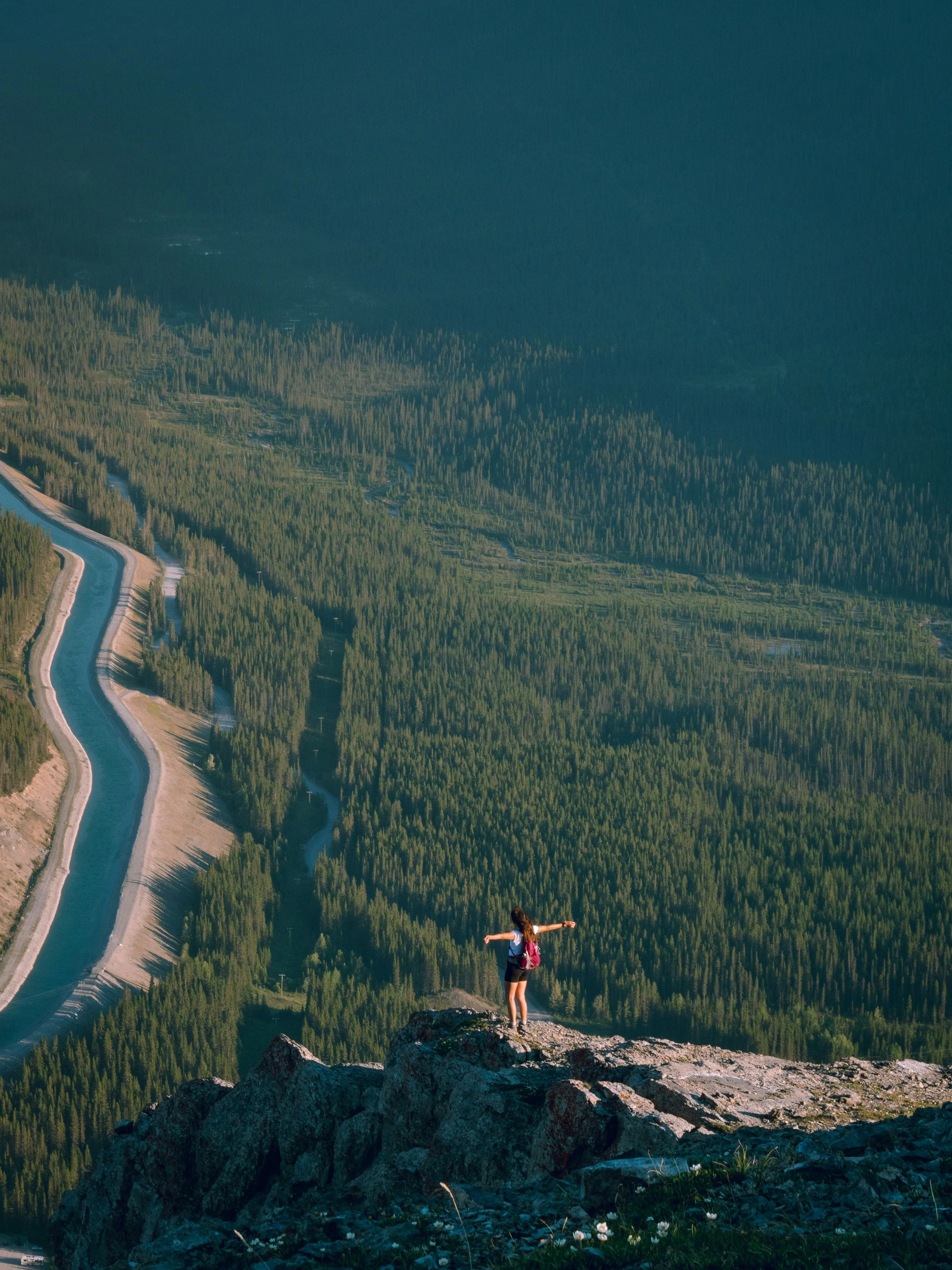 A person standing on a rocky ledge with arms outstretched, overlooking a vast forested landscape with a winding road below.