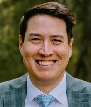 Smiling man outdoors wearing a suit and tie with a blurred green background.